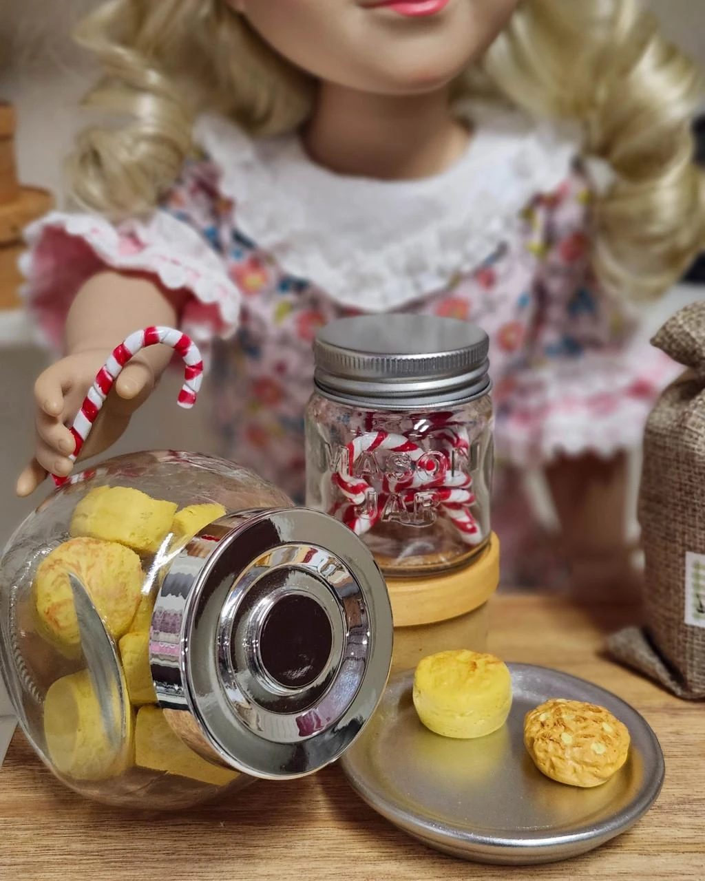 Sized for a 18" American Girl® Doll holds a tiny candy cane next to miniature c jars filled with doll-sized biscuits, cookies, and peppermint candy canes on a wooden counter