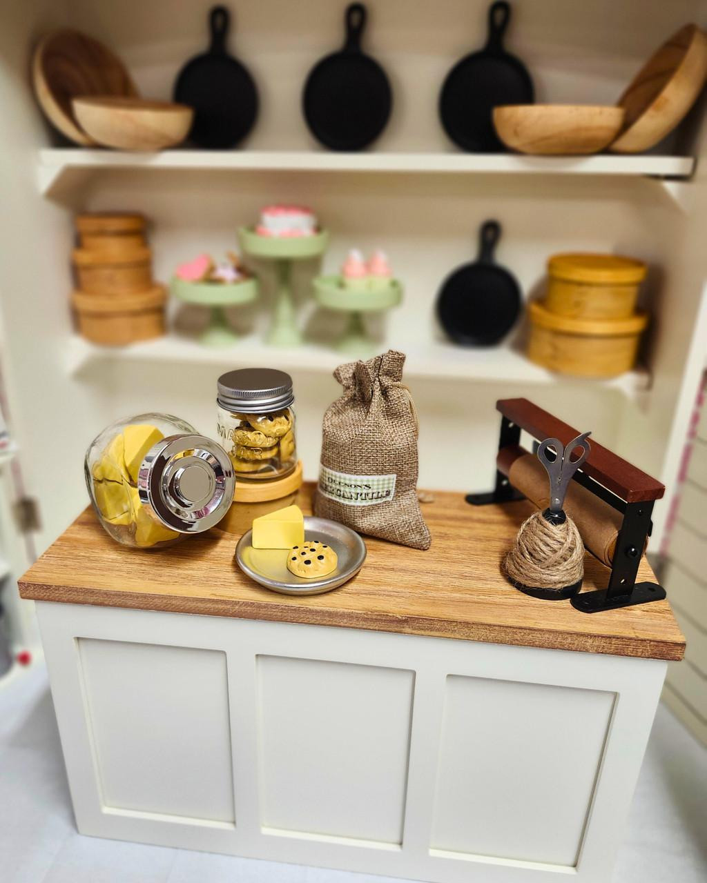 A miniature wooden store counter and shelving display by The Queens Treasures®, stocked with miniature cookie jars, a burlap sack, tiny plates of treats, cast-iron skillets, and a wooden twine dispenser.