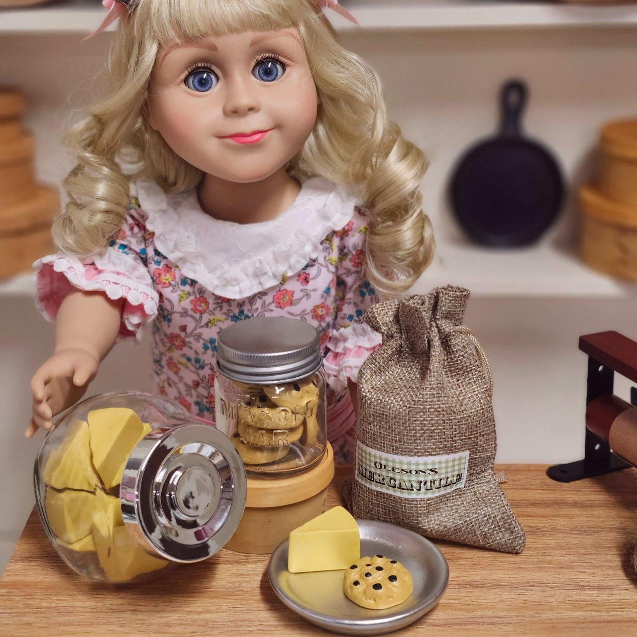 An 18"  Doll stands behind a wooden counter displaying miniature store goods, including a clear jar filled with a cookie jar, a small burlap sack, and a tiny plate with cheese and a cookie sized for 18" American Girl® dolls.