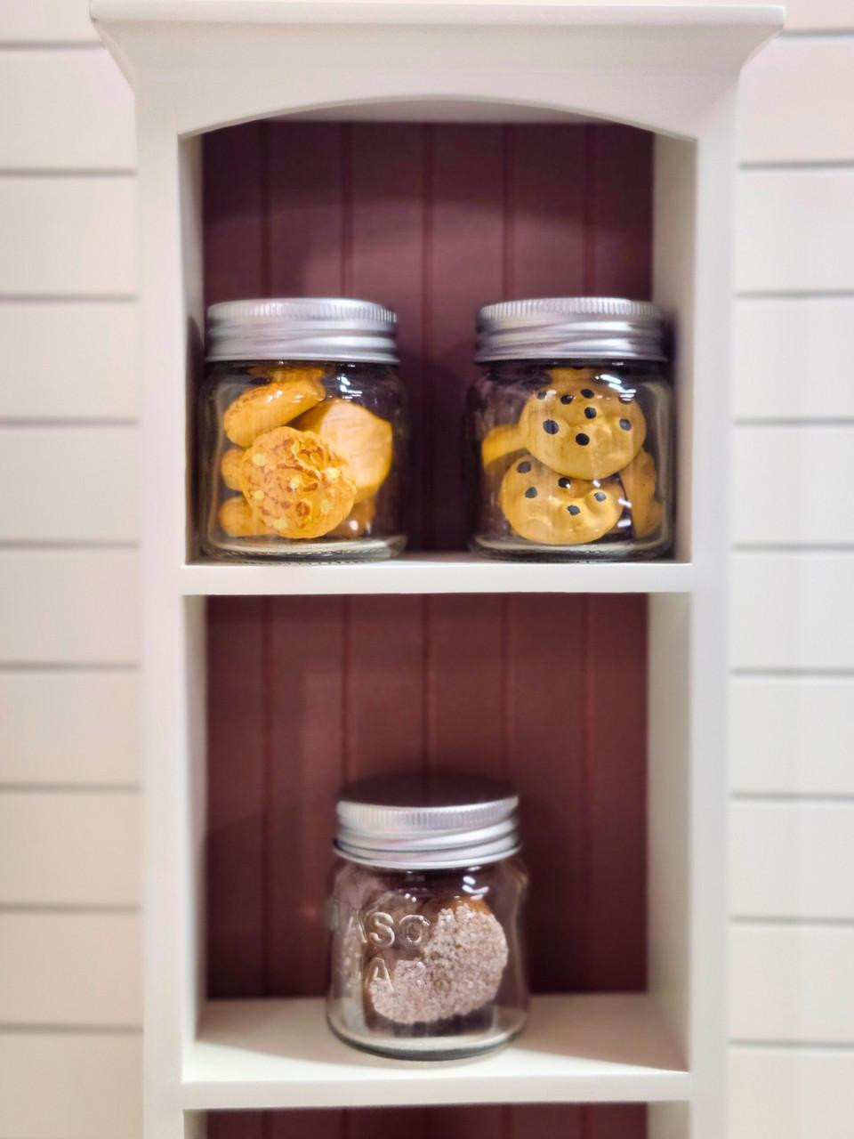 Three miniature clear cookie jars with silver lids, filled with tiny baked treats scaled for 18" American Girl® Dolls, displayed on the shelves of a white and reddish-brown wooden cabinet unit.