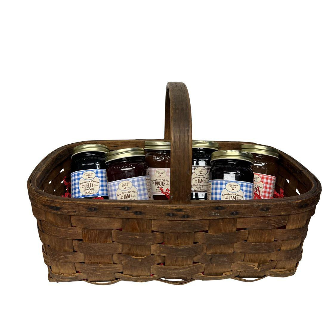 A rectangular dark woven basket with a single wooden handle, filled with an assortment of six Caroline's Kitchen jam jars featuring colorful gingham labels, displayed against a solid white background.