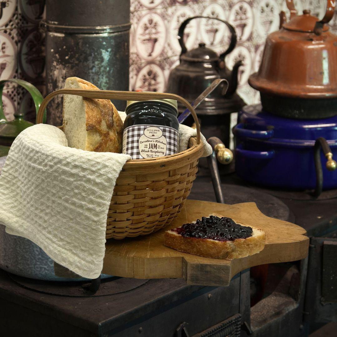 A jar of Caroline's Kitchen Black Raspberry Jam sits on a uniquely shaped pig wooden cutting board next to a slice of crusty bread and a slice spread with dark jam, displayed against a solid white background.