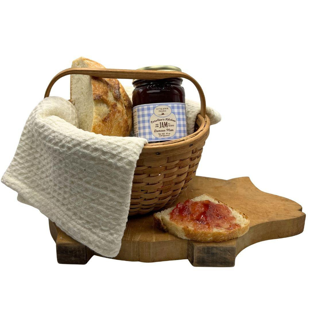 A jar of Caroline's Kitchen Damson Plum Jam nestled in a woven basket next to fresh bread, sitting on a wooden cutting board with a slice spread with jam, displayed against a solid white background.