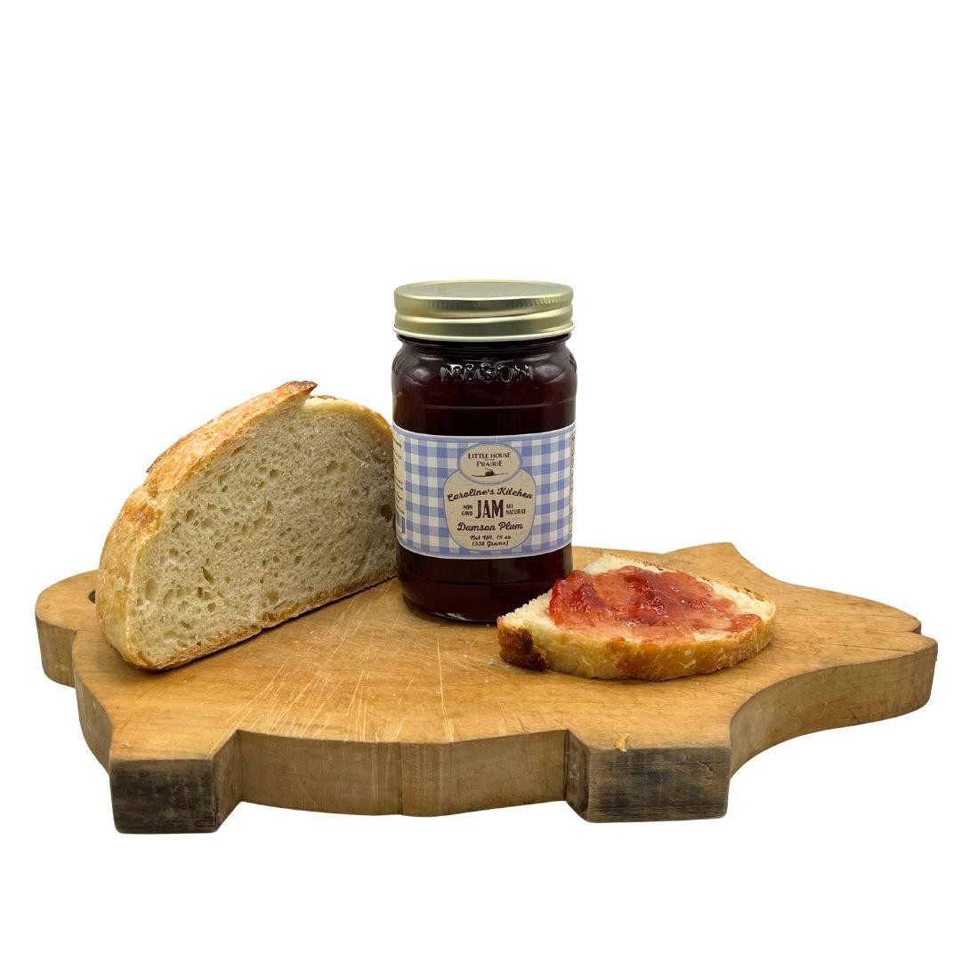 A jar of Caroline's Kitchen Damson Plum Jam sits on a uniquely shaped pig wooden cutting board next to a slice of crusty bread and a slice spread with red plum jam, displayed against a solid white background.