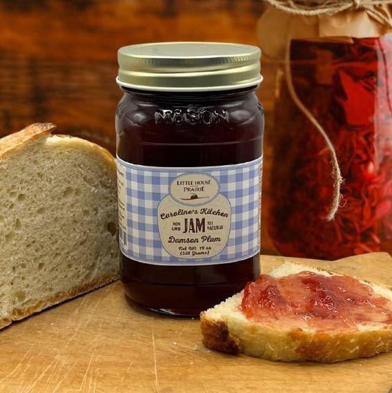 A jar of Caroline's Kitchen Damson Plum Jam with a light blue gingham label sits on a wooden board next to a loaf of crusty bread and a slice spread with plum jam.