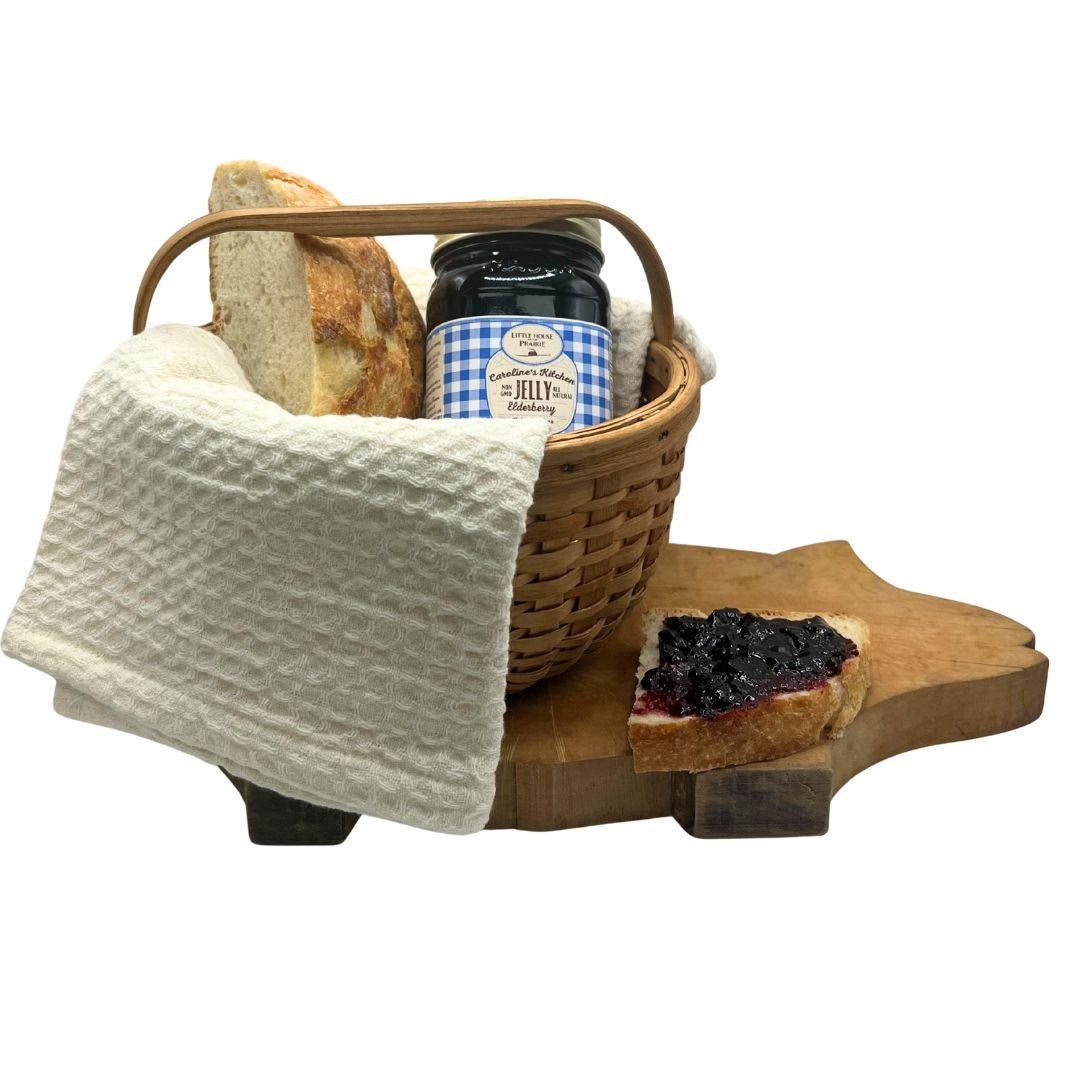 A jar of Caroline's Kitchen Elderberry Jelly nestled in a woven basket next to fresh bread, sitting on a uniquely shaped pig wooden cutting board with a slice spread with dark jelly, displayed against a solid white background.