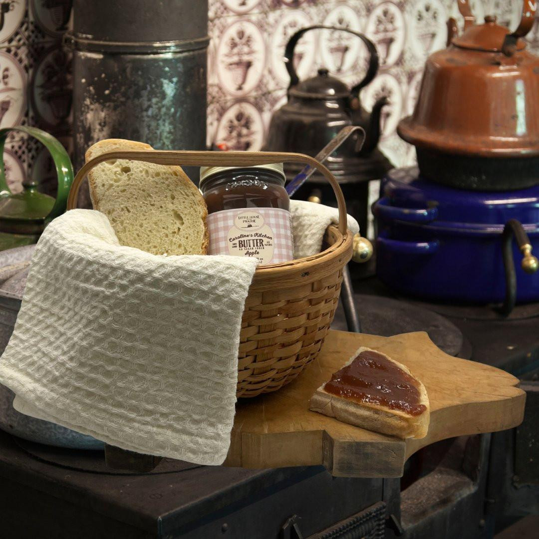 A jar of Caroline's Kitchen Apple Butter nestled in a woven basket next to fresh bread, sitting on a wooden cutting board with a slice spread with dark apple butter in front of vintage kitchen kettles.