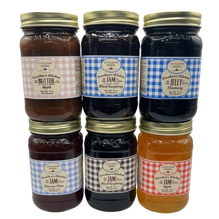 Six jars of Caroline's Kitchen fruit spreads arranged in two stacked rows, featuring flavors like Apple Butter, Black Raspberry Jam, Elderberry Jelly, and Danson Plum Jam, each with a distinctive gingham pattern label against a solid white background.