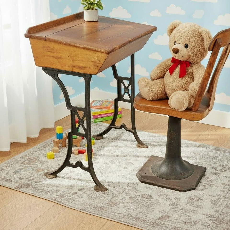 A vintage children's school desk and chair set sit on a patterned area rug in a room with blue sky and cloud wallpaper. The desk features a slanted wooden flip-top compartment supported by an ornate, dark cast-iron frame. Next to it, a matching wooden swivel chair holds a large, fluffy tan teddy bear wearing a red bow. Colorful toy blocks and books are visible on the floor in the background.