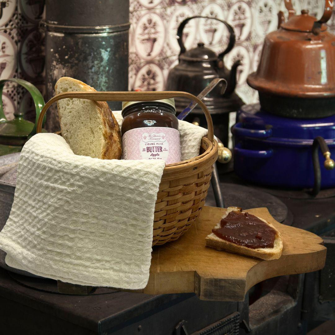 19 oz glass Mason jar of The Queen's Treasures Caramel Pecan Apple Butter placed in a basket with bread and a towel in an old fashiped room.
