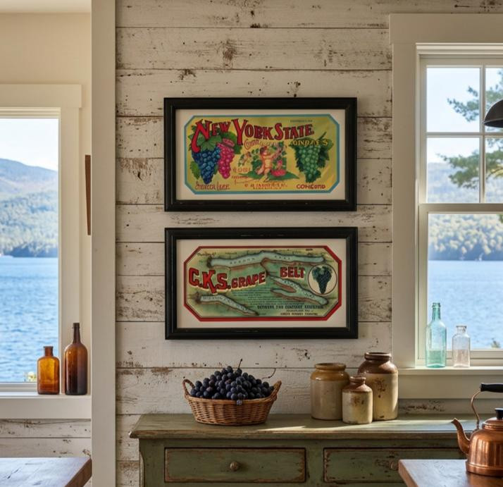 Framed Finger Lakes grape crate labels displayed on a whitewashed wood wall above a sideboard with fresh Concord grapes and stoneware crocks overlooking a lake
