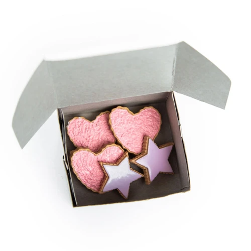 An open white cardboard bakery box is filled with an assortment of toy cookies against a plain white background. Inside the box are three heart-shaped cookies covered in pink sprinkles and two star-shaped cookies iced in smooth light purple.