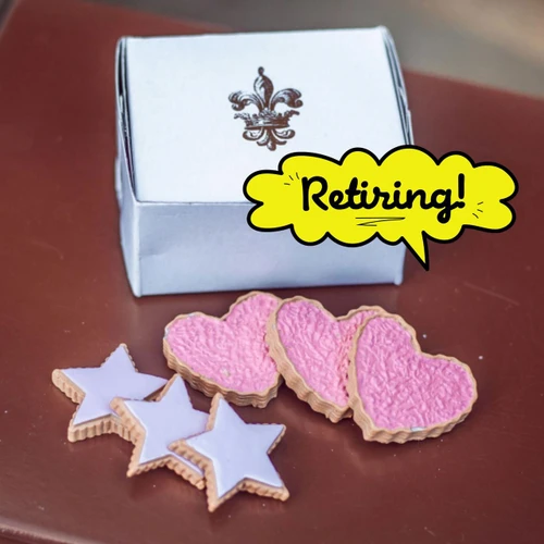 Three heart-shaped and three star-shaped frosted toy cookies sit on a dark surface in front of a white rectangular box with a crest logo. The heart cookies are frosted in pink sprinkles, while the star cookies are covered in solid light purple icing. A bright yellow speech bubble graphic points toward the box with the handwritten text "Retiring!"