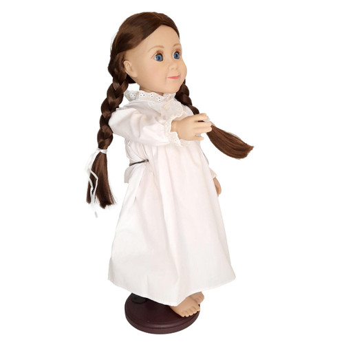 An 18-inch doll with brown hair styled in two long braids stands on a dark wooden display base against a solid white background. She is turned slightly to the side, wearing a long-sleeved, vintage-style white nightgown with a ruffled collar and cuffs.