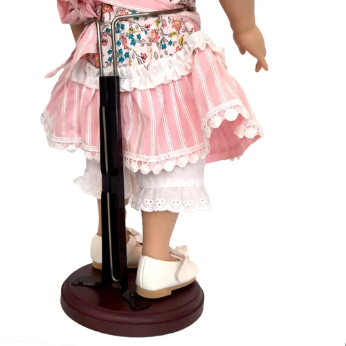 A close-up back view of an 18-inch doll secured in a dark brown wooden display stand against a solid white background. The adjustable silver metal waist wire wraps around the doll's torso under her pink and white striped dress, which features a floral apron overlay and white ruffled bloomers underneath.