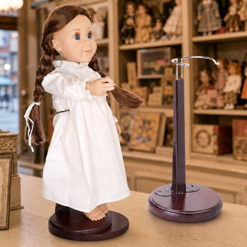 An 18-inch doll with brown hair styled in two long braids stands on a dark wooden display base against a solid white background. She is turned slightly to the side, wearing a long-sleeved, vintage-style white nightgown with a ruffled collar and cuffs. There is an empty doll stand. Both are in a store from the 1900's