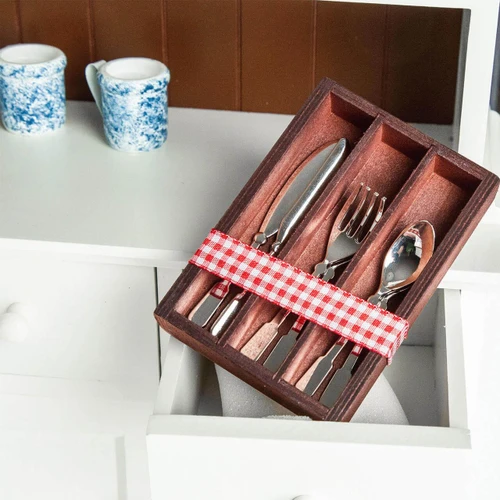 A set of doll-sized silverware designed to fit 18-inch American Girl® dolls is displayed in a wooden organizer tray, resting inside an open drawer of a white play kitchen. The dark reddish-brown wooden tray contains four mini metal butter knives, four forks, and four spoons divided into slots, all secured together by a red and white checkered ribbon. Two small, blue-and-white patterned ceramic mugs sit on the counter space in the background.