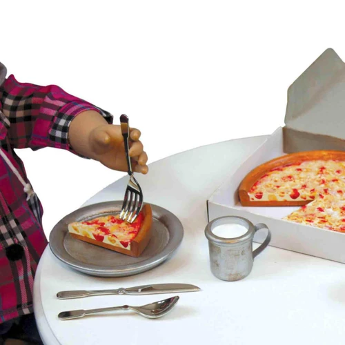 A close-up shot focuses on an 18-inch doll's hand holding a miniature metal fork, pressing it into a slice of toy pizza on a small grey plate. The scene is set on a white table, which also holds a miniature metal knife, spoon, and a small metal mug filled with a white liquid resembling milk. In the background, a larger toy pizza sits inside an open white cardboard pizza box.