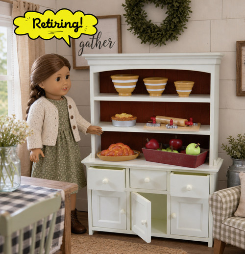 A doll-sized white wooden farmhouse cupboard and dish hutch made by The Queen's Treasures® is shown against a plain white background. The hutch features a dark brown interior backing with three open shelves holding miniature yellow nesting bowls, a small toy pie, a rolling pin with dough, a wooden plate of carrots, and a red crate filled with apples. The bottom buffet section has three drawers and three cabinets with small white knobs, with the two outer drawers and the center cabinet door slightly open. A bright yellow, comic-book-style speech bubble in the top left corner reads "Retiring!".