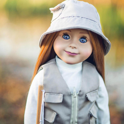 A close-up portrait of an 18-inch doll with long red hair and bright blue eyes, dressed in an outdoor fishing outfit. The doll wears a tan bucket hat with a stitched brim, a tan utility vest with pocket flaps, and a white high-neck shirt underneath. The background is a soft, warm-toned blur of orange and green natural scenery.