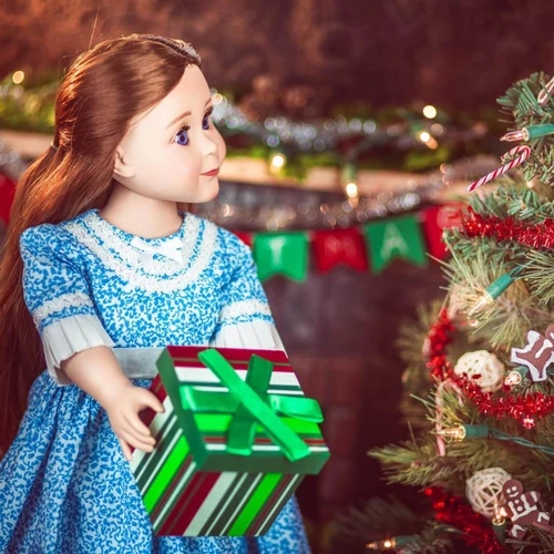 An 18-inch doll with long brown hair stands next to a decorated Christmas tree, holding a miniature green-and-red striped gift box with a green bow. The doll is wearing a blue-and-white patterned holiday dress with lace trim. In the background, a warm fireplace mantel is decorated with festive green and red banner flags and blurry holiday garland lights.