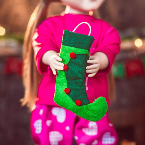 An 18-inch doll wearing bright pink pajamas with a white pattern holds a miniature elf-style Christmas stocking. The stocking is made of dark and light green felt with an upturned toe, a white loop for hanging, and a row of small red pom-poms decorating the front seam. The background is a softly blurred holiday setting with festive lights.