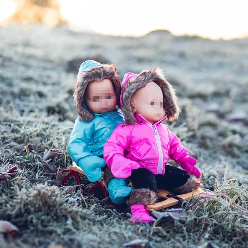 Two baby dolls ride together on a small wooden sled down a frosty, grassy slope outdoors. The doll in front wears a bright pink hooded winter coat with black pants, matching pink boots, and mittens, while the doll behind wears a matching light blue winter coat outfit.