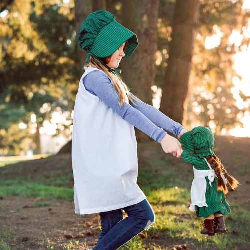 A girl wearing the child size little house on the prairie apron and bonnet sits with an 18 inch doll wearing matching 18" doll clothing outfit. A girl wearing the child size little house on the prairie apron and bonnet sits with an 18 inch doll wearing matching 18" doll clothing outfit.