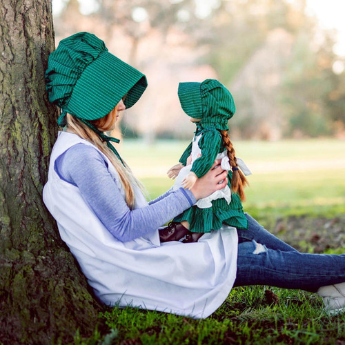 A girl wearing the child size little house on the prairie apron and bonnet sits with an 18 inch doll wearing matching 18" doll clothing outfit. A girl wearing the child size little house on the prairie apron and bonnet sits with an 18 inch doll wearing matching 18" doll clothing outfit.
