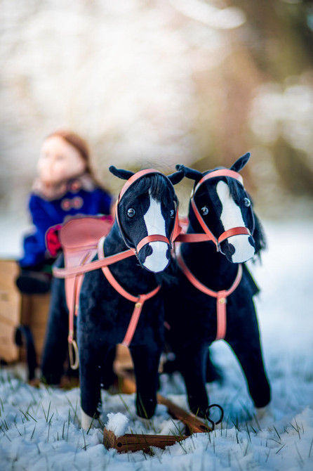 A close-up view shows two toy black horses with white facial markings wearing red harnesses as they stand in a snowy field, with a blurred 18-inch doll sitting in a wooden sleigh in the background.