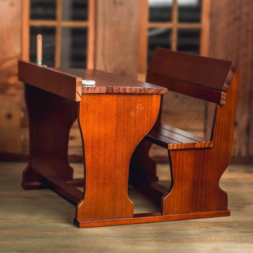 A miniature wooden pioneer school desk with an attached bench set inside a log cabin room.