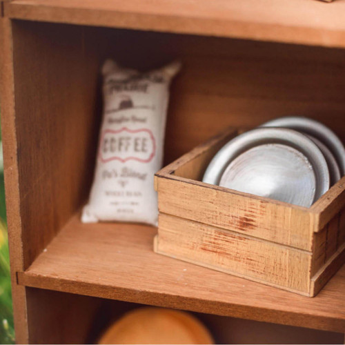 A close-up view of a wooden doll storage shelf holding a miniature fabric sack of coffee beans next to three small metal plates stacked inside a tiny wooden crate.