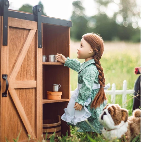 An 18-inch pioneer doll with a long brown braid stands outdoors next to a small plush dog, reaching into the shelves of a wooden barn-door storage trunk.