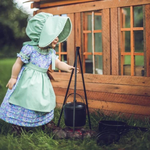 In outdoor play scene, an 18 inch doll dressed in our purple summer dress set reaches over the cookfire.