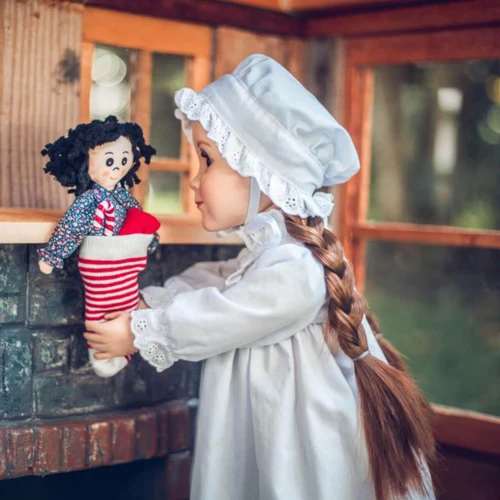 In holiday scene, an 18 inch doll dressed in Little House on the Prairie Night Gown reaches for her Christmas stocking hanging above the hearth.