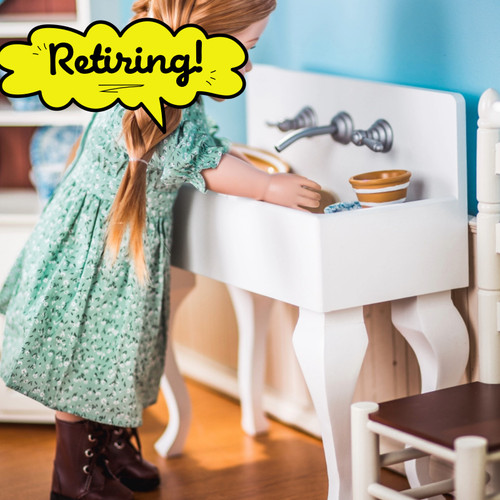 An 18-inch doll with long blonde braids stands at a white wooden vintage-style kitchen sink by The Queen’s Treasures®. She wears a green floral dress and appears to be washing miniature plates and bowls in the deep basin, which features a silver faux faucet with two cross handles against a light blue wall background.