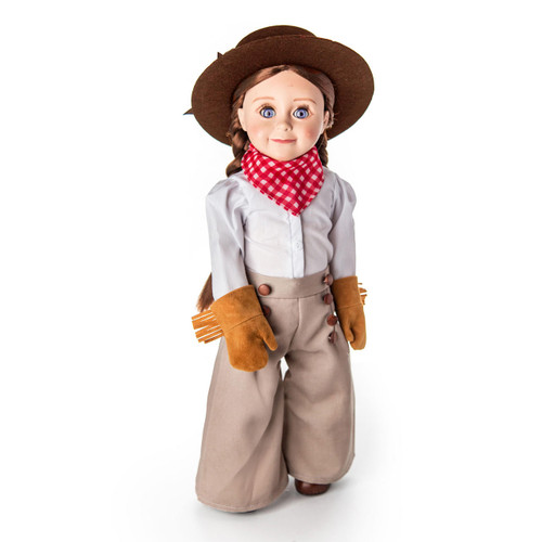 An 18-inch doll with blue eyes and light brown hair styled in low pigtails stands facing forward against a plain white background. The doll is wearing a historical frontier or cowboy outfit consisting of a light grey long-sleeved shirt, wide-legged tan trousers with button details, tan fringe gloves, a red and white checkered neck bandana, and a brown wide-brimmed cowboy hat.