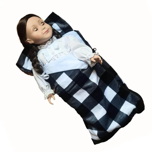 This studio product photograph shows an 18-inch doll tucked into a miniature plaid sleeping bag, isolated against a solid white background.