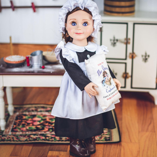 An 18-inch doll with brown hair stands facing forward in a miniature vintage kitchen setting, holding a small white play sack of flour. She is dressed in a historic maid uniform featuring a black long-sleeved dress, a ruffled white apron, and a matching white mop cap. The background shows a white wooden table with a dark top to the left and a white vintage-style toy icebox to the right, all resting on a light wood floor with a patterned rug.