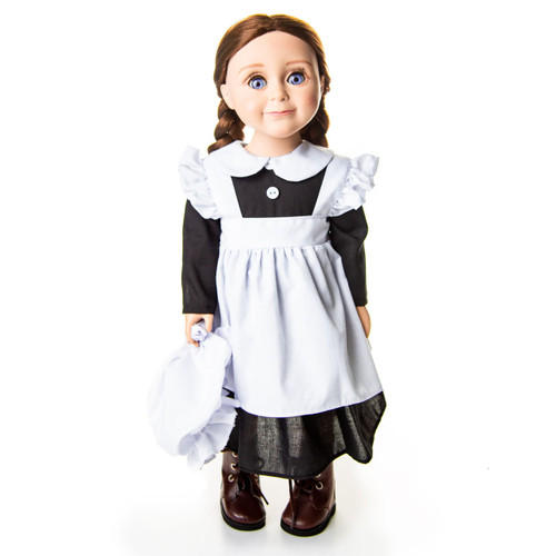 An 18-inch doll with brown hair in two low braided pigtails stands facing forward against a plain white background. She is wearing a historic kitchen maid outfit consisting of a black long-sleeved dress, a crisp white pinafore apron with ruffled shoulder straps, and dark brown leather-look boots. She holds a matching white fabric mop cap in her right hand down by her side.