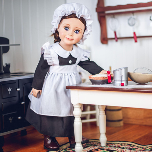An 18-inch doll with brown hair stands in a miniature vintage kitchen wearing a historic maid uniform that fits American Girl® dolls. She is dressed in a black long-sleeved dress, a ruffled white apron, and a white mop cap. The doll stands behind a white wooden table with a dark top, reaching toward miniature kitchen play accessories, including a mixing bowl and a metal can. A vintage black toy stove is visible on the left, and a wooden utensil rack hangs on the white-paneled wall in the background.