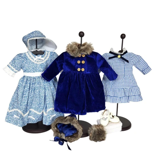 An assortment of three historical-style 18-inch doll outfits and matching accessories are displayed on black stands against a plain white background. Left outfit: A light blue and white floral dress with white trim along the neckline, waist, and hem, complete with a matching ruffled bonnet. Center outfit: A royal blue velvet winter coat with gold double-breasted buttons and a brown faux-fur collar. A matching blue velvet bonnet with a faux-fur trim sits on the floor below it. Right outfit: A light blue and white gingham-patterned dress featuring a white collar, a black ribbon bow at the neckline, and a ruffled hem. Accessories: A pair of white doll shoes and a small brown faux-fur muff sit on the floor to the right