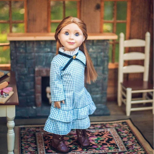 An 18-inch doll  wearing a light blue and white gingham-patterned long-sleeved dress with a ruffled skirt, a white lace collar, and a black ribbon tie at the neck. She stands on a patterned area rug in front of a gray stone fireplace, with a wooden chair visible to her right