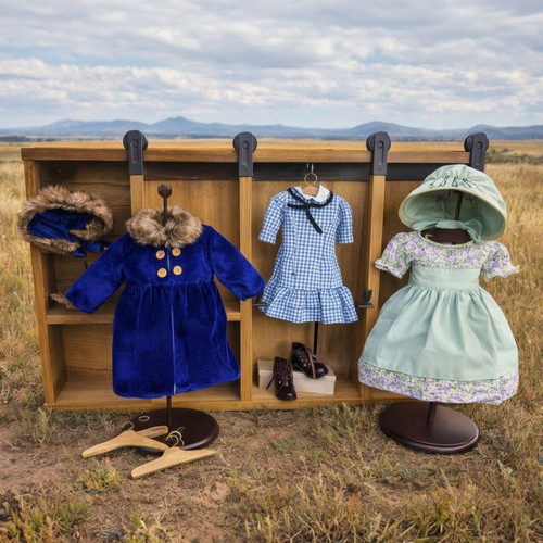 18 inch doll trunk with barn style sliding doors to reveal different compartments, red checkered dress, blue velvet coat, purple summer dress and brown lace up boots placed in a prairie outdoor setting