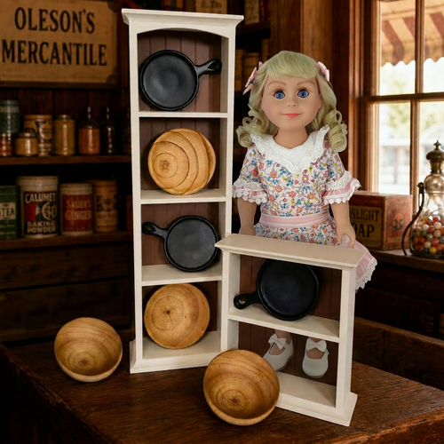 image shows 3 bowls and 2 frying pans in cream colored shelving displayed in a store setting With our Nellie Oleson 18inch doll behind the shelves