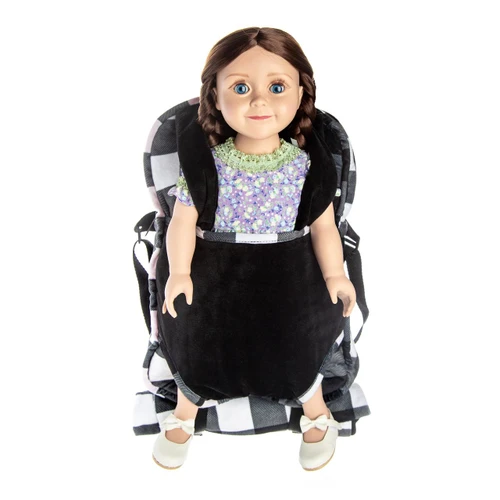 A doll is secured facing forward in a plush doll-carrier backpack against a plain white background. The backpack features a solid black front pouch and straps layered over a black and white buffalo plaid main bag.