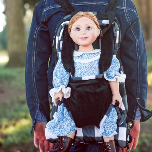 A doll with light brown hair and blue eyes is strapped into a black doll-carrier backpack featuring black and white plaid accents. The backpack is worn by a person in a blue denim jacket. A matching plaid rolled-up sleeping bag is visible at the bottom of the carrier.