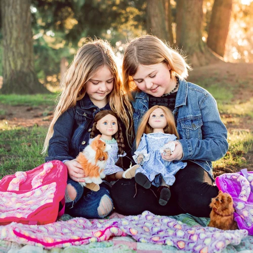 Two girls sit on a blanket outdoors, each holding an 18 inch doll and, with patterned backpacks doll carriers beside them.