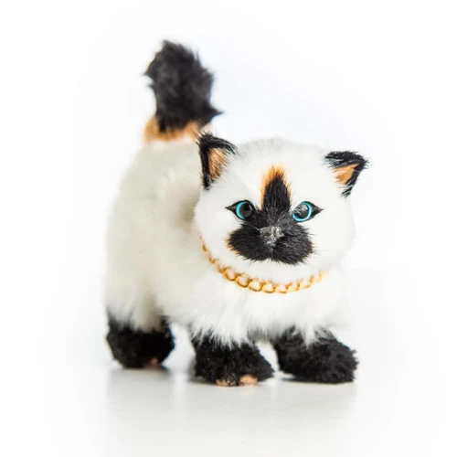 A realistic, fluffy Siamese-style toy cat stands facing forward on a plain white background. The toy cat features a cream-colored body with dark brown ears, paws, and face mask, along with light blue eyes and a small patch of tan fur at the base of its upright black tail. It wears a polished gold link chain collar around its neck.