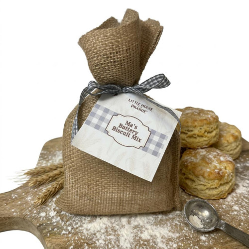 A rustic burlap sack of Ma's Buttery Biscuit Mix tied with a gingham ribbon sits on a floured wooden cutting board next to fresh biscuits, wheat stalks, and a metal measuring spoon against a white background.